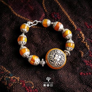 Puede incluir: Una pulsera con cuentas redondas de color naranja transl&uacute;cido que se alternan con intrincados espaciadores de metal plateado. Un gran dije central circular de metal plateado con un patr&oacute;n floral est&aacute; integrado al dise&ntilde;o.