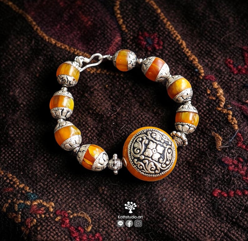 Puede incluir: Una pulsera con cuentas redondas de color naranja transl&uacute;cido que se alternan con intrincados espaciadores de metal plateado. Un gran dije central circular de metal plateado con un patr&oacute;n floral est&aacute; integrado al dise&ntilde;o.