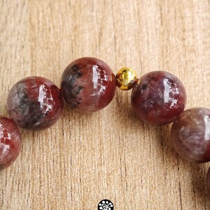 Peut inclure: Un bracelet de perles compos&eacute; de pierres sph&eacute;riques polies aux tons rouge fonc&eacute; et noir marbr&eacute;, s&eacute;par&eacute;es par une petite perle m&eacute;tallique dor&eacute;e et r&eacute;fl&eacute;chissante. Le logo Kathstudio.art est imprim&eacute; en bas avec des ic&ocirc;nes de r&eacute;seaux sociaux.