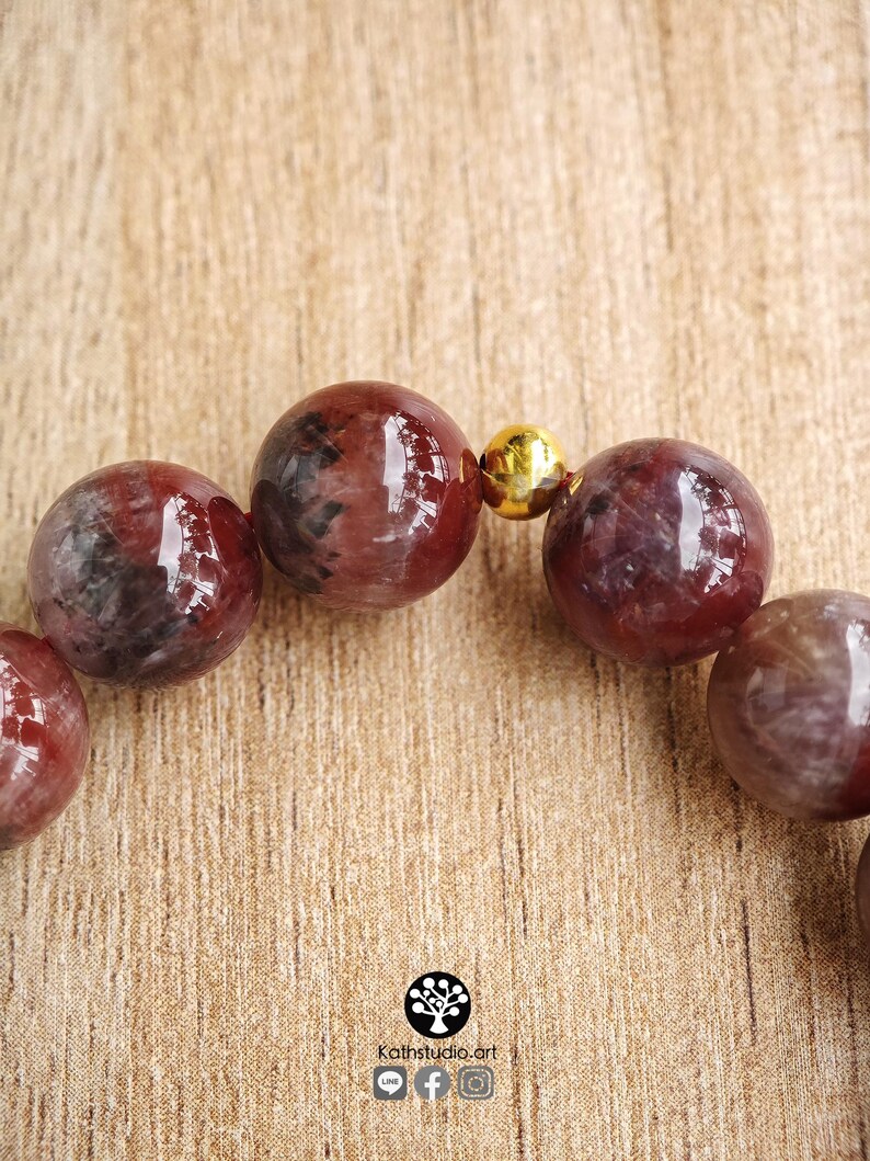 Peut inclure: Un bracelet de perles compos&eacute; de pierres sph&eacute;riques polies aux tons rouge fonc&eacute; et noir marbr&eacute;, s&eacute;par&eacute;es par une petite perle m&eacute;tallique dor&eacute;e et r&eacute;fl&eacute;chissante. Le logo Kathstudio.art est imprim&eacute; en bas avec des ic&ocirc;nes de r&eacute;seaux sociaux.