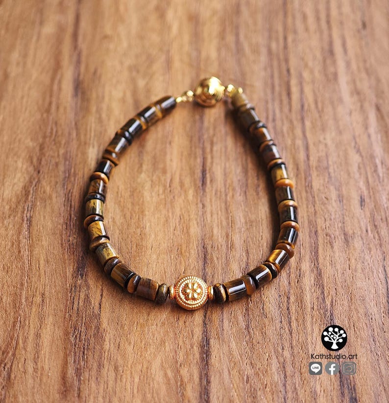 May include: A bracelet composed of cylindrical tiger's eye beads in shades of brown and gold, featuring a central round gold-toned metal bead with a floral embossed design and a gold-toned spherical clasp closure at the top of the circular arrangement.
