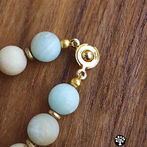 May include: A beaded bracelet featuring matte light blue and off-white spherical beads separated by small gold-toned metal spacers. The bracelet includes a circular gold-toned metal magnetic clasp mechanism. The text Kathstudio.art is visible.