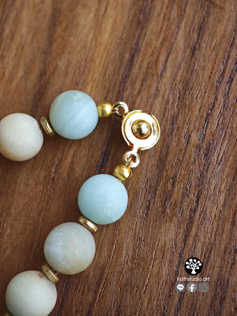 May include: A beaded bracelet featuring matte light blue and off-white spherical beads separated by small gold-toned metal spacers. The bracelet includes a circular gold-toned metal magnetic clasp mechanism. The text Kathstudio.art is visible.