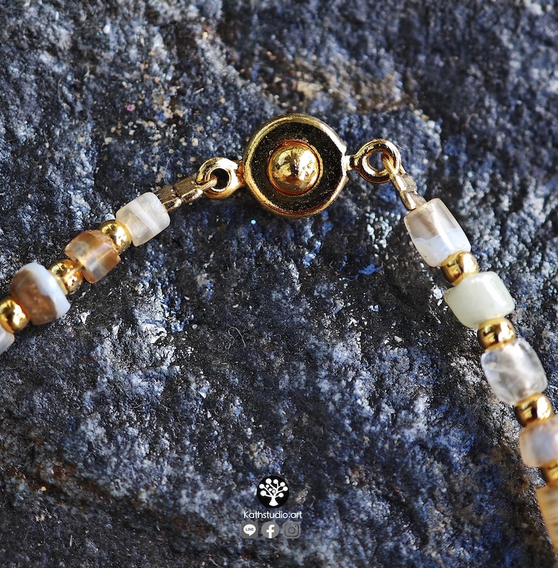 May include: A beaded bracelet featuring alternating translucent, earth-toned stone beads and small gold-colored metallic spheres. The bracelet is secured with a circular gold-colored magnetic clasp. The text Kathstudio.art is visible at the bottom.