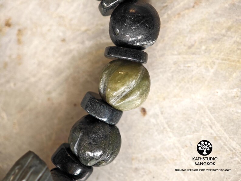 Puede incluir: Una pulsera de cuentas que alterna cuentas negras redondas y cuentas esf&eacute;ricas de color verde oliva talladas. Las cuentas negras est&aacute;n intercaladas con espaciadores negros finos y planos. La imagen incluye el texto KATHSTUDIO BANGKOK TURNING HERITAGE INTO EVERYDAY ELEGANCE.