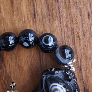 May include: A bracelet featuring polished black agate beads with white and grey banded eye patterns. The beads are strung together, and one bead is attached to a carved black stone pendant with a silver-toned metal cap. The text Kathstudio.art is visible.