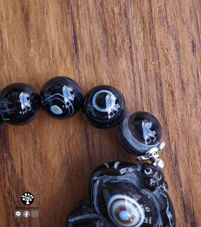 May include: A bracelet featuring polished black agate beads with white and grey banded eye patterns. The beads are strung together, and one bead is attached to a carved black stone pendant with a silver-toned metal cap. The text Kathstudio.art is visible.