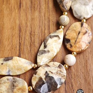 Puede incluir: Un collar de varias hebras con cuentas de piedra pulidas de formas irregulares en tonos beige y marr&oacute;n, intercaladas con peque&ntilde;as cuentas espaciadoras redondas de metal dorado. Las piedras muestran patrones naturales de veteado.