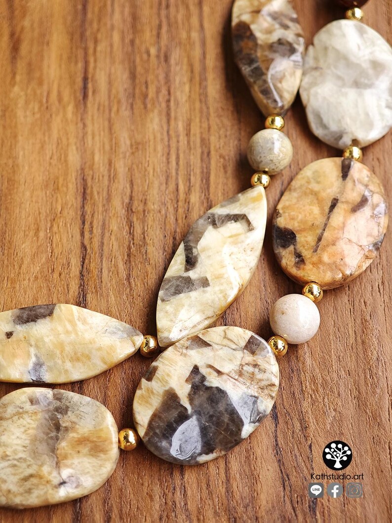 Puede incluir: Un collar de varias hebras con cuentas de piedra pulidas de formas irregulares en tonos beige y marr&oacute;n, intercaladas con peque&ntilde;as cuentas espaciadoras redondas de metal dorado. Las piedras muestran patrones naturales de veteado.
