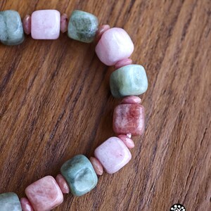 May include: A beaded bracelet featuring alternating square-shaped polished stones in shades of pale pink, dusty rose, and sage green, separated by small, round, pink spacer beads. The bracelet is arranged in a curved line on a wooden surface.
