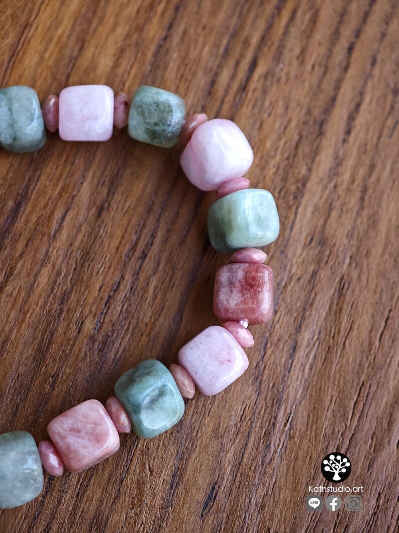 May include: A beaded bracelet featuring alternating square-shaped polished stones in shades of pale pink, dusty rose, and sage green, separated by small, round, pink spacer beads. The bracelet is arranged in a curved line on a wooden surface.
