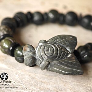 Puede incluir: Una pulsera con un dije central de cigarra tallado en piedra, flanqueado por cuentas esf&eacute;ricas alternas en verde oscuro y negro. La cigarra presenta patrones de alas grabados y un ojo prominente. El texto KATHSTUDIO BANGKOK es visible.