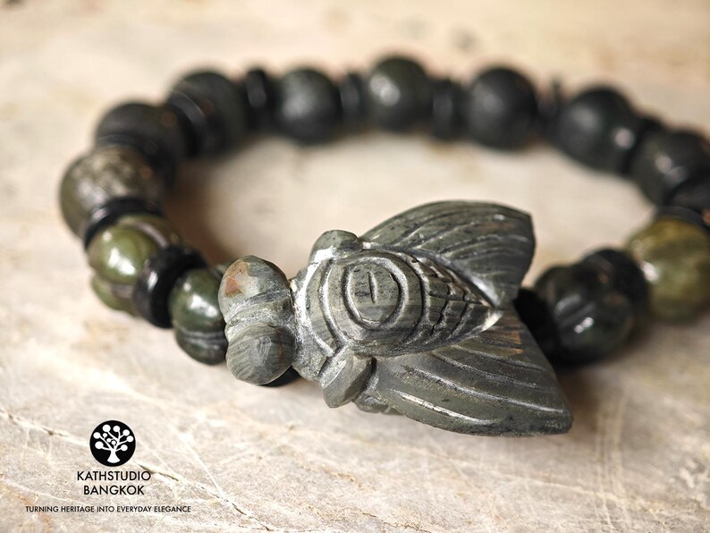 Puede incluir: Una pulsera con un dije central de cigarra tallado en piedra, flanqueado por cuentas esf&eacute;ricas alternas en verde oscuro y negro. La cigarra presenta patrones de alas grabados y un ojo prominente. El texto KATHSTUDIO BANGKOK es visible.