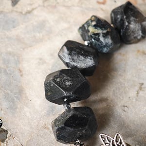 May include: A bracelet featuring large, faceted, dark gray to black stones with irregular shapes. Small, metallic, dark-colored spacer beads are placed between the stones. A silver-toned metal charm with an intricate, openwork design is attached.