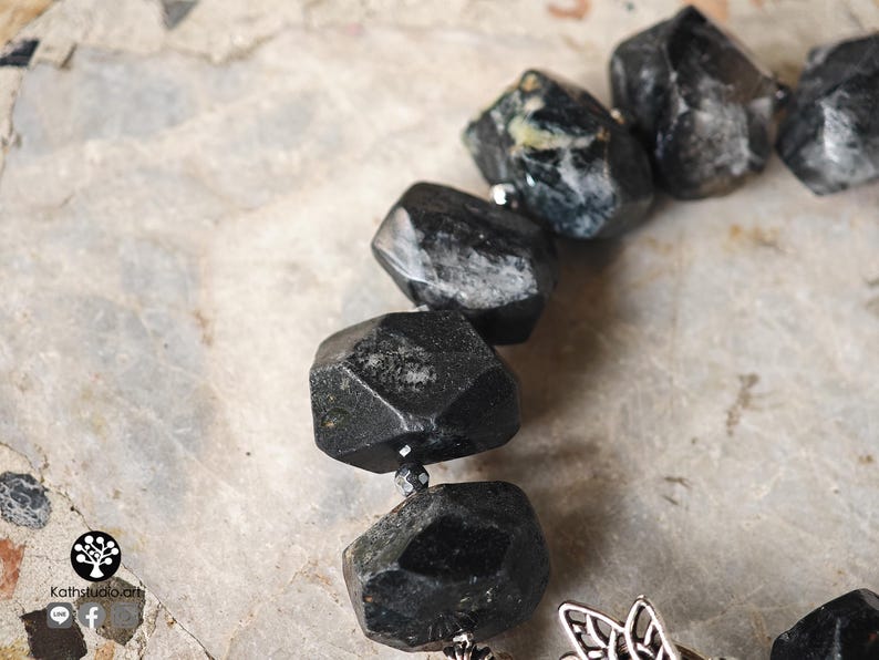 May include: A bracelet featuring large, faceted, dark gray to black stones with irregular shapes. Small, metallic, dark-colored spacer beads are placed between the stones. A silver-toned metal charm with an intricate, openwork design is attached.