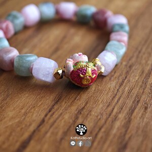 May include: A beaded bracelet featuring alternating square-cut beads in shades of pale pink, soft green, and translucent white. The centerpiece is a gold-toned charm with a red and green painted finish, flanked by small gold-colored metallic spacers.