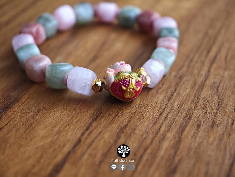 May include: A beaded bracelet featuring alternating square-cut beads in shades of pale pink, soft green, and translucent white. The centerpiece is a gold-toned charm with a red and green painted finish, flanked by small gold-colored metallic spacers.