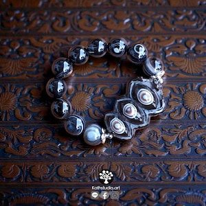 May include: A beaded bracelet featuring round, dark, translucent beads with white and grey swirling patterns. A larger, central, irregular-shaped dark bead displays three distinct, concentric eye-like patterns in shades of brown and white.