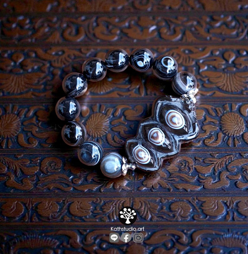 May include: A beaded bracelet featuring round, dark, translucent beads with white and grey swirling patterns. A larger, central, irregular-shaped dark bead displays three distinct, concentric eye-like patterns in shades of brown and white.