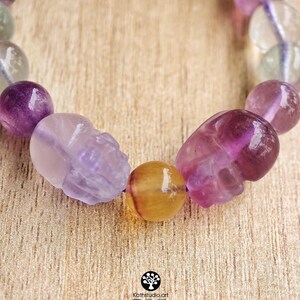 May include: A beaded bracelet featuring polished spherical beads in shades of purple, green, and clear, interspersed with two carved skull-shaped beads and a single yellow bead. The text Kathstudio.art is printed at the bottom with social icons.