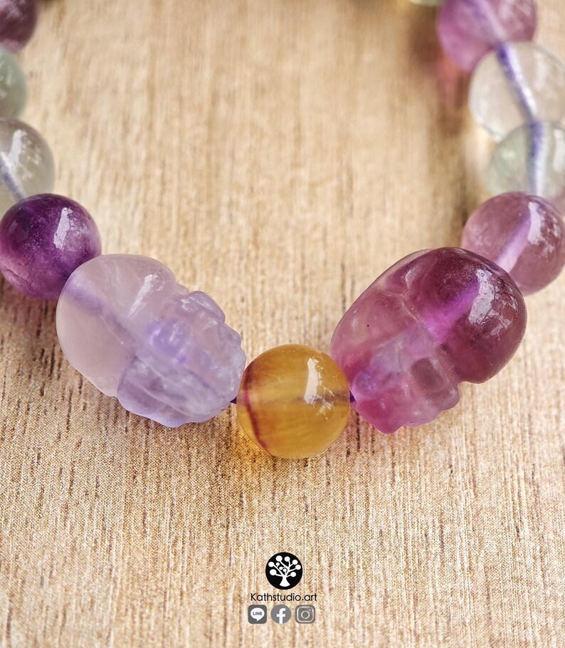 May include: A beaded bracelet featuring polished spherical beads in shades of purple, green, and clear, interspersed with two carved skull-shaped beads and a single yellow bead. The text Kathstudio.art is printed at the bottom with social icons.