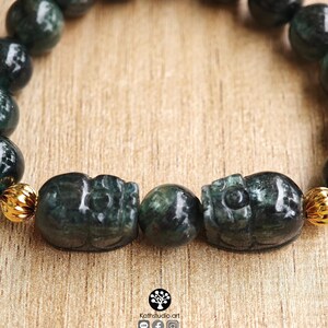 May include: A beaded bracelet featuring dark green, polished, spherical stone beads and two carved, dark green stone skull-shaped beads. The bracelet includes small, ribbed, gold-toned metal spacers between the beads. The logo Kathstudio.art is visible.