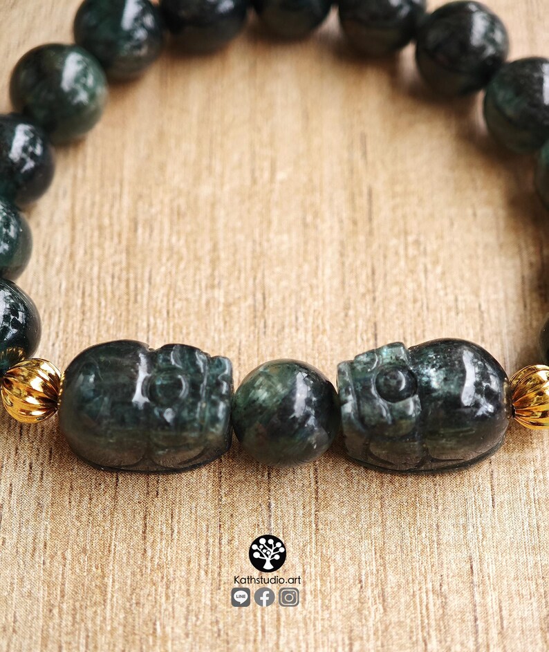May include: A beaded bracelet featuring dark green, polished, spherical stone beads and two carved, dark green stone skull-shaped beads. The bracelet includes small, ribbed, gold-toned metal spacers between the beads. The logo Kathstudio.art is visible.