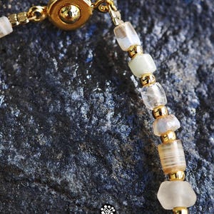May include: A beaded bracelet featuring translucent, off-white, and light brown cylindrical beads interspersed with small, polished gold-tone metal spacer beads. The bracelet includes a gold-tone circular magnetic clasp and a small logo graphic.