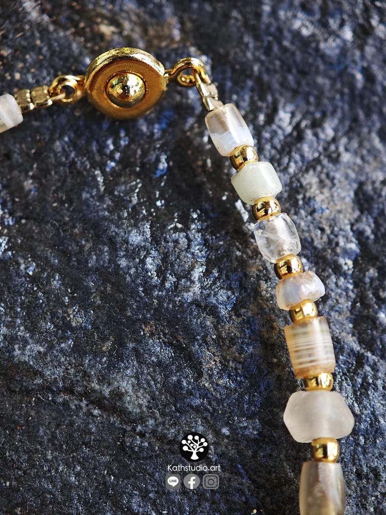 May include: A beaded bracelet featuring translucent, off-white, and light brown cylindrical beads interspersed with small, polished gold-tone metal spacer beads. The bracelet includes a gold-tone circular magnetic clasp and a small logo graphic.