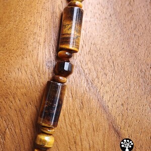 Puede incluir: Un collar con cuentas de ojo de tigre que alternan formas cil&iacute;ndricas y facetadas. Las piedras muestran patrones naturales de bandas de color marr&oacute;n dorado y marr&oacute;n oscuro con un acabado pulido. El texto Kathstudio.art es visible en la esquina.