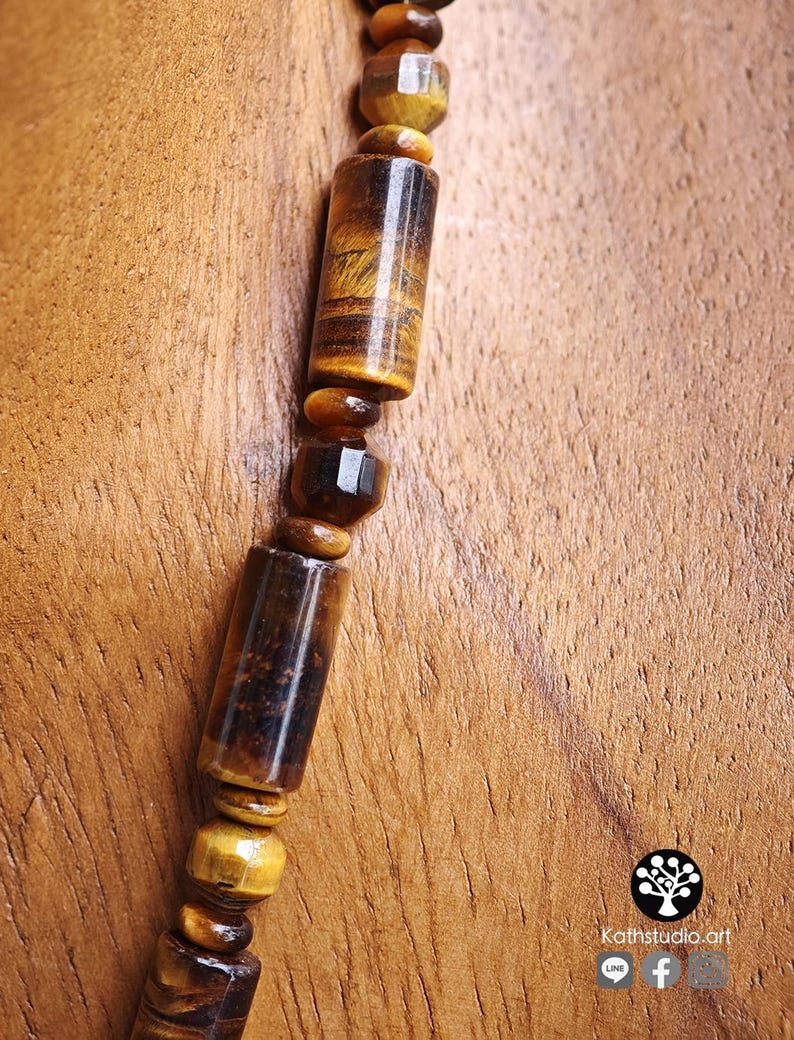 Puede incluir: Un collar con cuentas de ojo de tigre que alternan formas cil&iacute;ndricas y facetadas. Las piedras muestran patrones naturales de bandas de color marr&oacute;n dorado y marr&oacute;n oscuro con un acabado pulido. El texto Kathstudio.art es visible en la esquina.