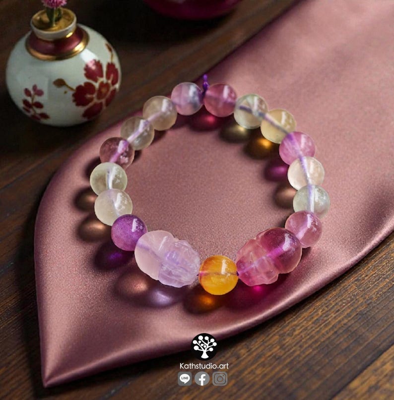May include: A beaded bracelet featuring translucent, multi-colored fluorite spheres in shades of purple, yellow, and pale green. The bracelet includes two carved skull-shaped beads and one smooth yellow bead, all strung on a purple cord.