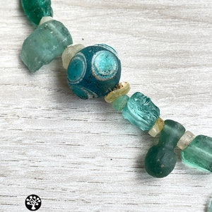 Puede incluir: Un collar con una cadena de cuentas de vidrio verde mar y turquesa de forma irregular. Una cuenta tiene una base azul oscuro con dise&ntilde;os circulares blancos y celeste. El collar se exhibe sobre una superficie de madera clara.
