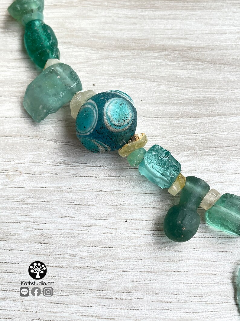 Puede incluir: Un collar con una cadena de cuentas de vidrio verde mar y turquesa de forma irregular. Una cuenta tiene una base azul oscuro con dise&ntilde;os circulares blancos y celeste. El collar se exhibe sobre una superficie de madera clara.