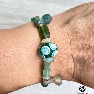 Puede incluir: Una pulsera hecha a mano con una variedad de cuentas de vidrio en tonos verdes, azules y transparentes. La pulsera incluye una cuenta grande de color verde oscuro con un dise&ntilde;o de remolino azul claro. Las cuentas est&aacute;n ensartadas con un cord&oacute;n resistente.