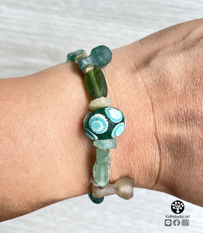 Puede incluir: Una pulsera hecha a mano con una variedad de cuentas de vidrio en tonos verdes, azules y transparentes. La pulsera incluye una cuenta grande de color verde oscuro con un dise&ntilde;o de remolino azul claro. Las cuentas est&aacute;n ensartadas con un cord&oacute;n resistente.