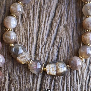 May include: A beaded bracelet featuring polished, translucent brown and grey moonstone beads interspersed with small gold-toned metal spacers. Two of the beads are carved into the shape of human skulls. The text Kathstudio.art is visible.