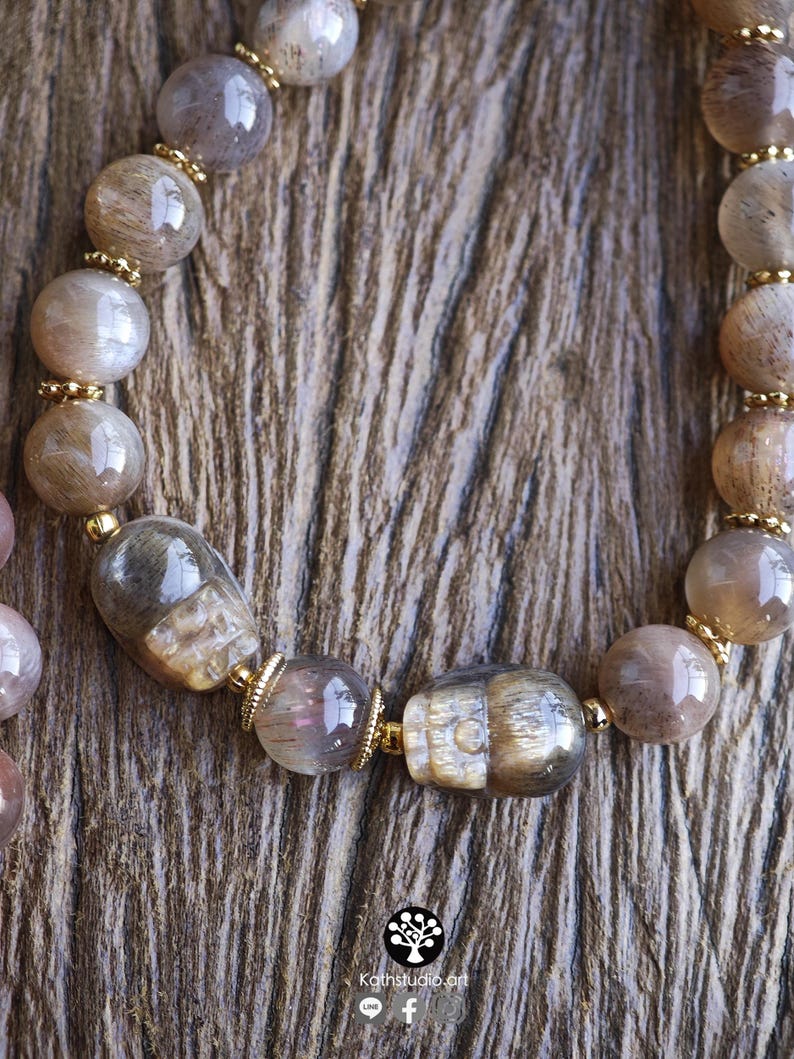 May include: A beaded bracelet featuring polished, translucent brown and grey moonstone beads interspersed with small gold-toned metal spacers. Two of the beads are carved into the shape of human skulls. The text Kathstudio.art is visible.