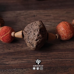May include: Close-up of a handcrafted necklace featuring a central, textured, brown stone bead flanked by orange and wooden beads. The necklace is set against a dark wooden surface, highlighting the natural tones and textures of the materials.