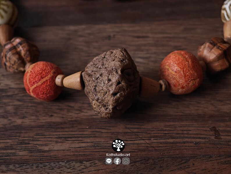 May include: Close-up of a handcrafted necklace featuring a central, textured, brown stone bead flanked by orange and wooden beads. The necklace is set against a dark wooden surface, highlighting the natural tones and textures of the materials.