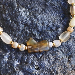 May include: A beaded bracelet featuring a central carved brown stone animal charm, interspersed with translucent off-white stone beads and small metallic gold-toned spacer beads, arranged in a repeating pattern along the length of the jewelry.