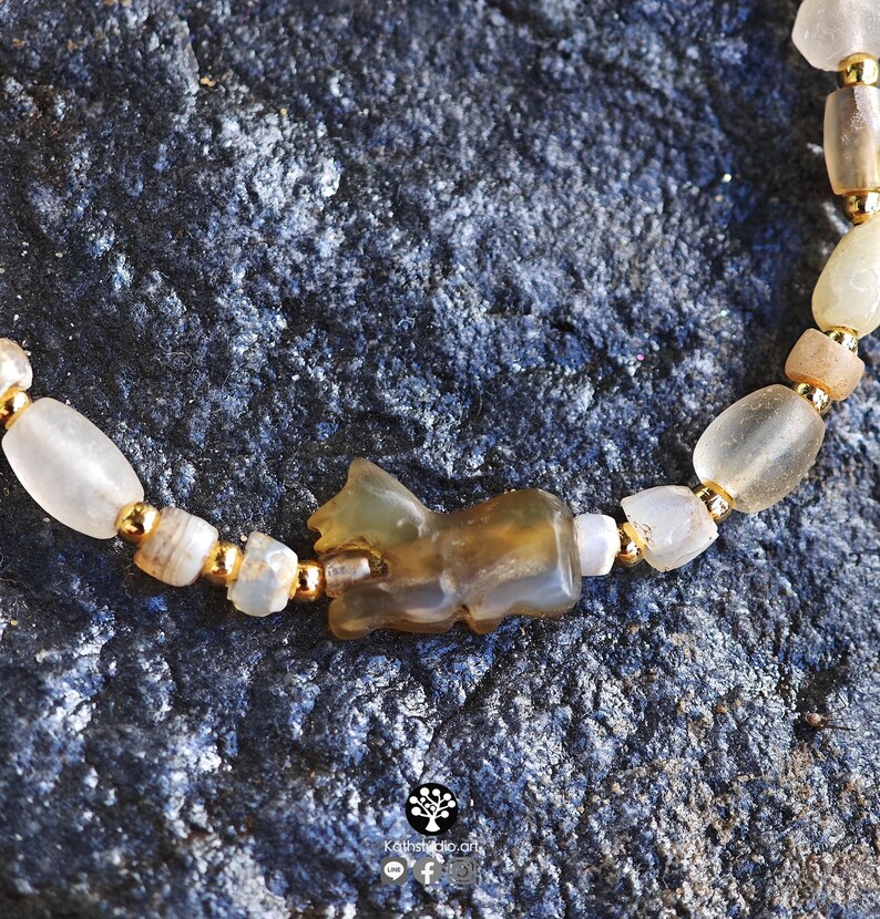 May include: A beaded bracelet featuring a central carved brown stone animal charm, interspersed with translucent off-white stone beads and small metallic gold-toned spacer beads, arranged in a repeating pattern along the length of the jewelry.