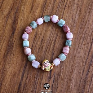 May include: A beaded bracelet featuring alternating square-cut beads in shades of pale pink, dusty rose, and sage green, accented by small gold-toned spacers and a central gold-toned charm depicting a stylized figure with a painted face.