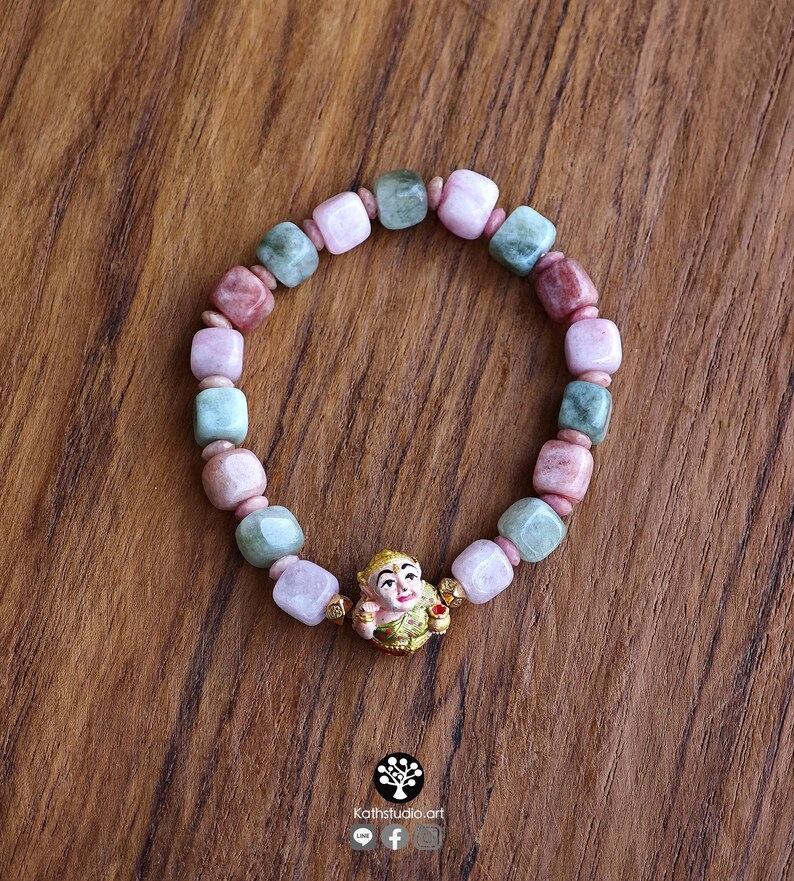 May include: A beaded bracelet featuring alternating square-cut beads in shades of pale pink, dusty rose, and sage green, accented by small gold-toned spacers and a central gold-toned charm depicting a stylized figure with a painted face.