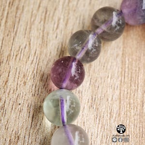 May include: A beaded bracelet featuring smooth, spherical fluorite beads in shades of translucent purple, grey, and pale green. The beads are strung together on a vibrant purple cord. A small logo with the text Kathstudio.art is visible.