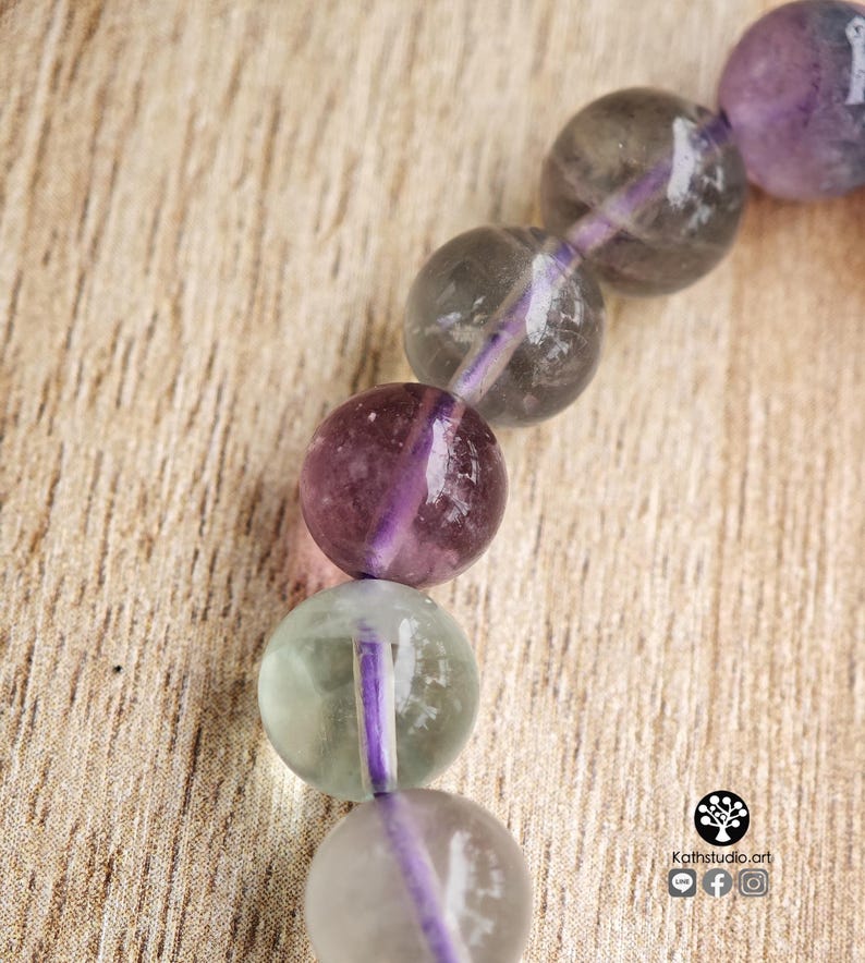 May include: A beaded bracelet featuring smooth, spherical fluorite beads in shades of translucent purple, grey, and pale green. The beads are strung together on a vibrant purple cord. A small logo with the text Kathstudio.art is visible.