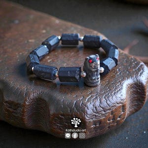May include: A bracelet composed of dark, cylindrical, faceted beads and small metallic spacers. The focal point is a carved, dark animal-shaped bead featuring two small, bright red eyes. The text Kathstudio.art is visible at the bottom.