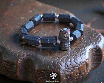 Op de afbeelding: Een armband bestaande uit donkere, cilindrische, gefacetteerde kralen en kleine metalen tussenstukjes. Het middelpunt is een uitgesneden, donkere, diervormige kraal met twee kleine, felrode ogen. De tekst Kathstudio.art is onderaan zichtbaar.