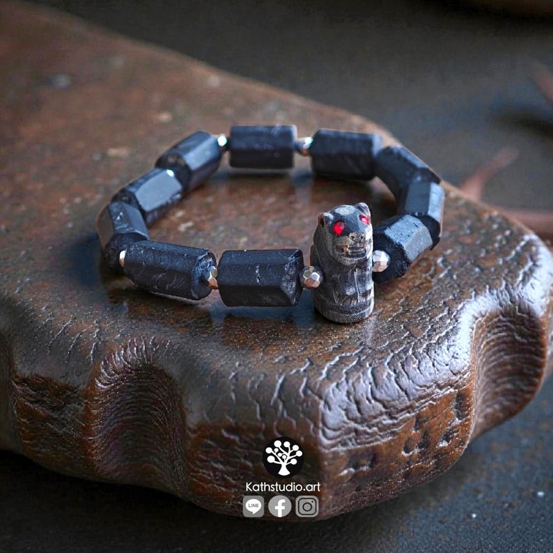 May include: A bracelet composed of dark, cylindrical, faceted beads and small metallic spacers. The focal point is a carved, dark animal-shaped bead featuring two small, bright red eyes. The text Kathstudio.art is visible at the bottom.