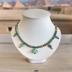 Op de afbeelding: Een turquoise kralenketting met kleine, vierkante kralen en verschillende turquoise bedels in de vorm van vogels en olifanten. De ketting wordt getoond op een witte buste met een vage achtergrond van piramides.